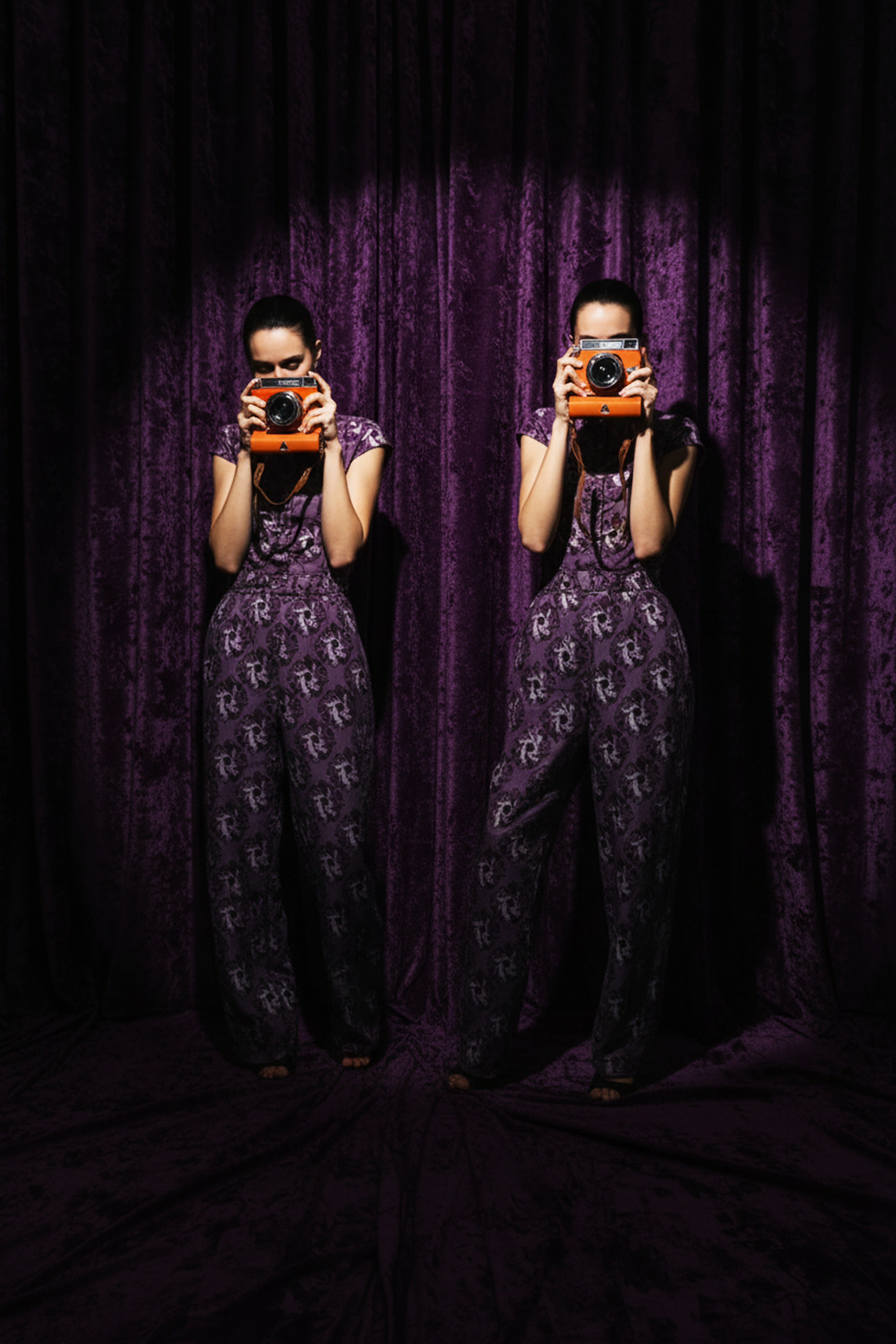 Person wearing a patterned outfit holding a camera in front of a purple curtain.