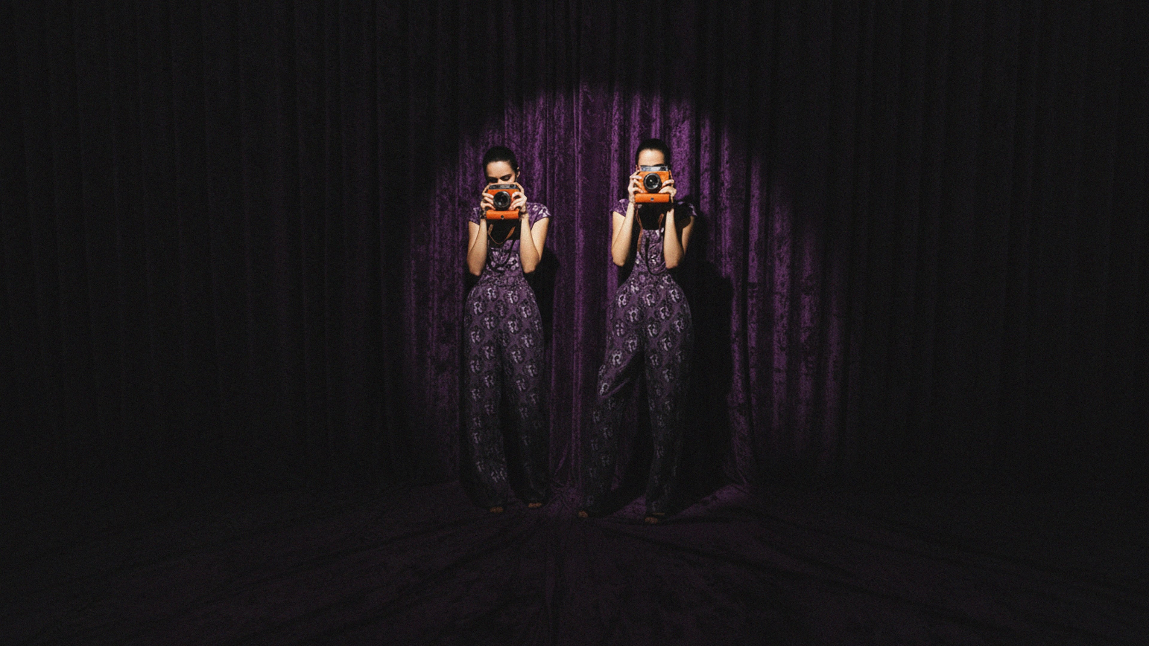 Two people in matching outfits standing in front of a purple curtain, holding cameras.