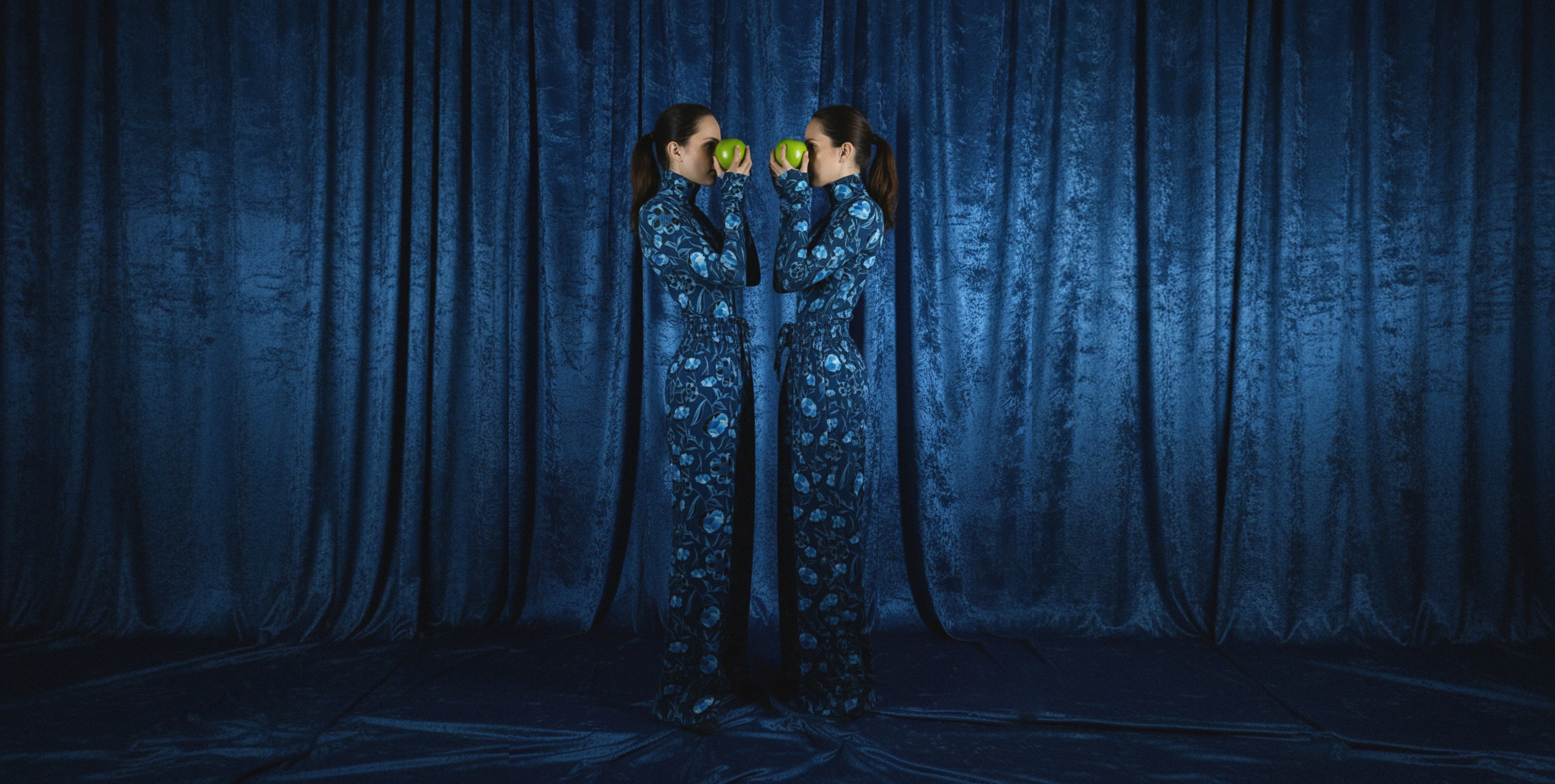Two people in matching outfits holding glowing green spheres against a dark curtain background